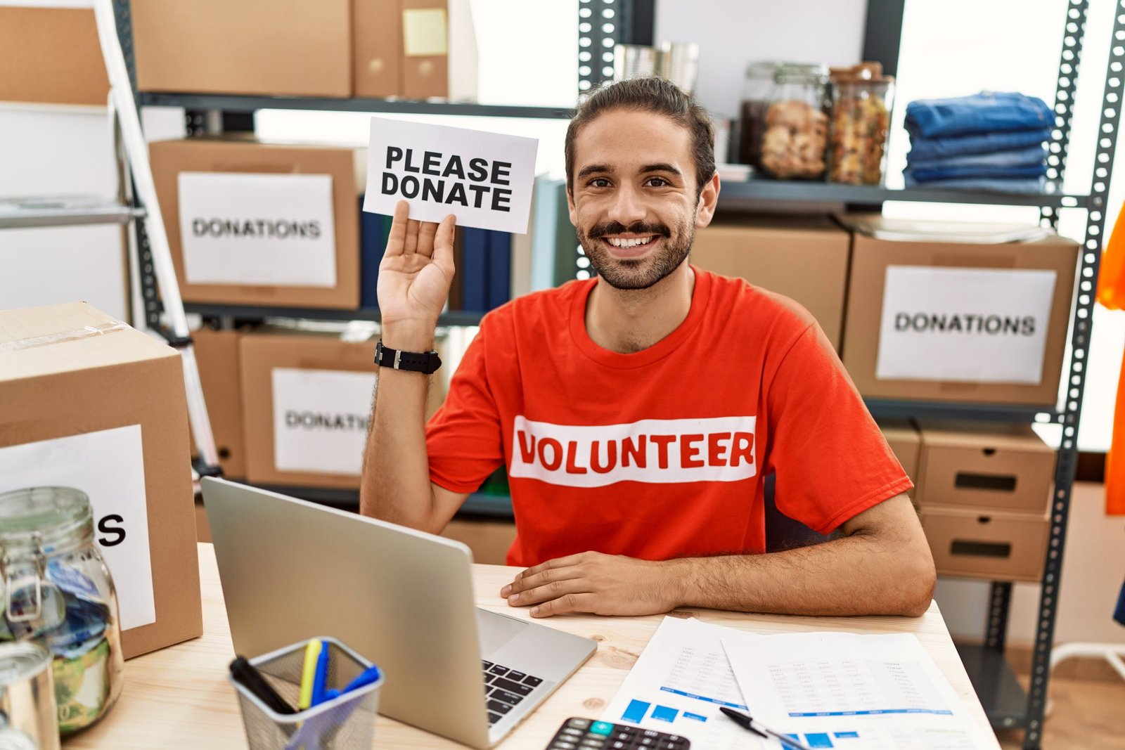 Home young hispanic man wearing volunteer t shirt holding please donate banner looking positive and happy standing and smiling with a confident smile showing teeth